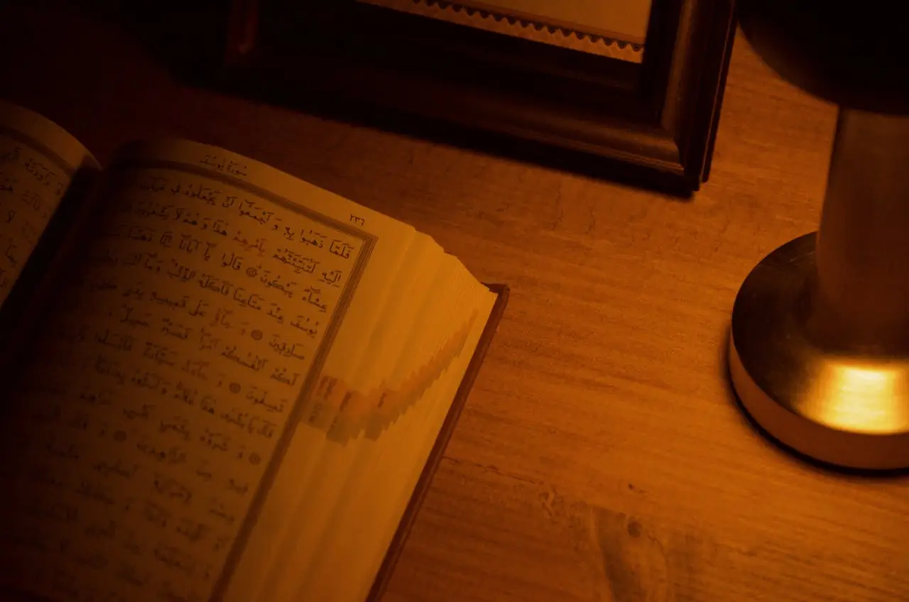about-01 An open Quran on a wooden table next to a lit lamp, set in warm ambient light.
